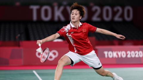 Chen Yu Fei lunges forward playing a shot. She is wearing a red collard shirt with white shorts and white trainers. She has short brown hair.