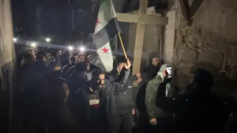 AFP Two men lead a crowd of people through a narrow street, one holding the Syrian rebel flag and another a large wooden cross.