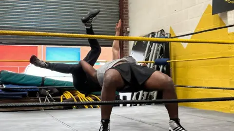 The character Lysander preparing to slam the character Demetrius to the ground inside a wrestling ring.