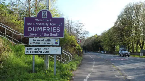Billy McCrorie A signpost next to a road saying "welcome to the University Town of Dumfries - Queen of the South"