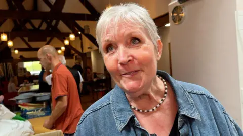 A woman with short white hair is wearing a denim jacket and a brown and white beaded necklace. She is inside a room that has wooden beams on the ceiling and handing lights. There are people behind her. 