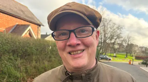 A man wearing a brown flat cap and a weathered, buttoned-up coat stands on a residential street lined with hedges and brick houses. He is wearing black glasses. Leafless trees and a small park area are visible in the background under a partly cloudy sky.