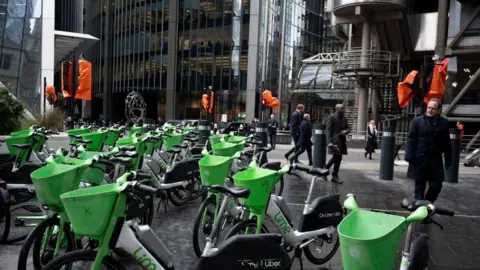 Rows of green and white Lime rental bikes are parked in Leadenhall in the City of London, with people walking by in professional attire. The background features modern glass office buildings, including the Lloyd's building. Several statues and poles are covered in orange fabric.