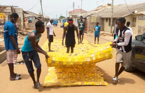 Nii Odzenma People rolling out yellow tapestry tapestry designed by artist Serge Attukwei Clottey on a road in La - Accra, Ghana