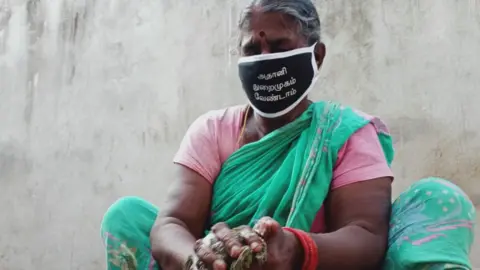 के सरवनन बंदरगाह विस्तार के विरोध में मास्क पहनकर एक महिला बर्तन धो रही है