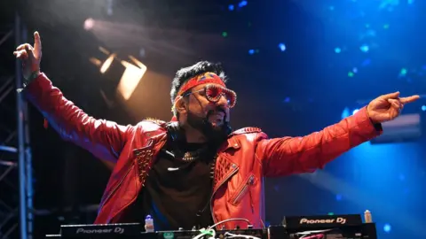 DJ Bobby Friction and the Bhangra All Stars perform on the Truth stage during day 2 of Glastonbury Festival 2023. Bobby is a British Asian man in his early 50s. He wears a studded red leather jacket over a black T-shirt paired with a red headband and red tinted sunglasses. His eyes are closed as he holds both arms out above his DJ deck, his head turned to the left. He's on stage, lit by spotlights and blue light
