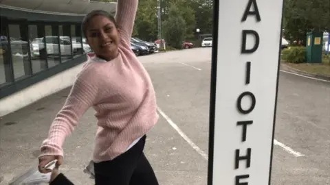 Sania Ahmed Sania is wearing a pink sweater and black trousers. She is jumping up smiling at the camera, with the sign to the hospital next to her. The background is of a car park, with glass buidlings on the left and cars directly behind, with trees and leaves overhanging.