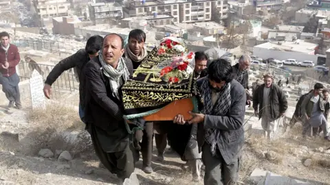 EPA a group of men carrying a coffin up a dusty hill, with city buildings in the distance