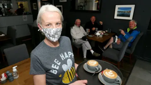 PAcemaker A waitress with blonde hair wearing a face mask and holding a tray with coffees on it looks at the camera, with a table with three men sitting at it behind her