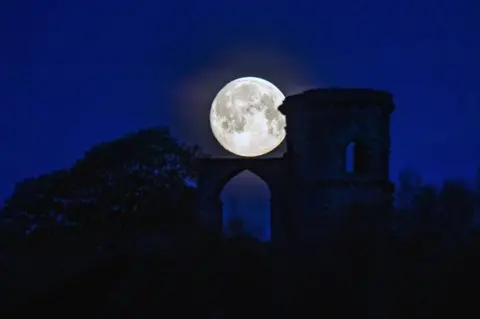 PA Media The pink supermoon seen against the silhouetted Mow Cop Folly in Cheshire, UK