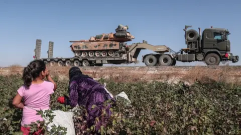 EPA Seasonal workers cut a cotton while Turkish military vehicles carrying tanks as they are on the way to Northern Syria for a military operation in Kurdish areas, near the Syrian border, near Akcakale district in Sanliurfa, Turkey 12 October 2019.