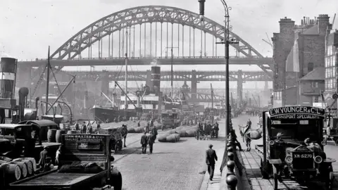 Newcastle Local Studies Library/Tyne Bridge Black and white view of vehicles on quayside with Tyne Bridge beyond