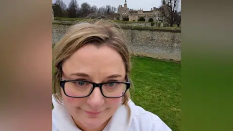 Rhian Williams Woman with fair hair and glasses stands in front of a stately home