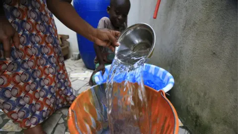 EPA A woman fills a bucket with tap water in Abidjan, Ivory Coast as a small boy watches on.