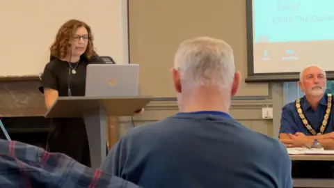 Councillor Polly Lamb speaking into a microphone while reading from her laptop in a council meeting