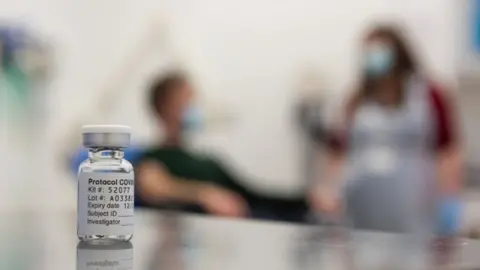 John Cairns/University of Oxford HO A test volunteer and medical worker behind a vial of the Oxford-AstraZeneca vaccine