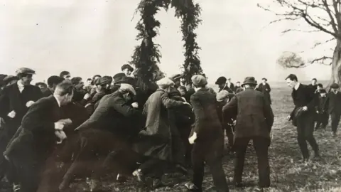 Alnwick Shrovetide Football Committee  Men wearing suits and caps surrounding the goalposts known as hales in the 1920s