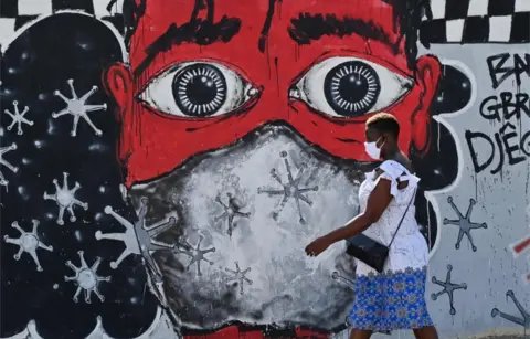 AFP A woman wearing a mask walks past a large mural depicting a person wearing a mask.