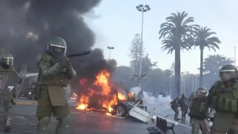 Riot police walk past a burning car