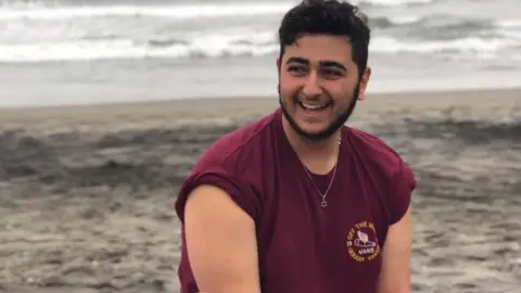 Ben Mizrachi family Young man with beard and red shirt sitting at the shore, water in background