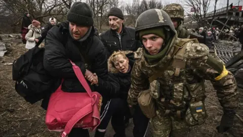 Getty Images A visibly exhausted woman is assisted while crossing a destroyed bridge in the city of Irpin, northwest of Kyiv