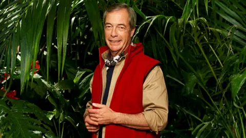 ITV/Shutterstock A man with short grey hair stands in front of a jungle background of dense foliage, made up of long, green leaves that hang down around him. He's standing with his hands clasped together, and wears a khaki (light brown) shirt with a red waistcoat/body warmer over the top. He's got a neckerchief on with a black, white and burgundy camouflage pattern.