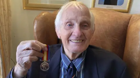 BBC David Elam. He has short white hair and blue eyes. He is sat on a brown leather chair and is wearing a blue shirt and a navy blue jacket and tie. He is holding his silver Nuclear Test Medal and smiling at the camera.