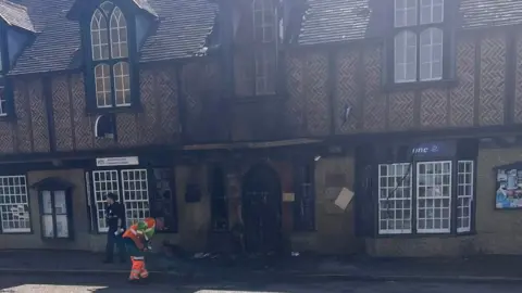 Nickita Starck A fire damaged timber-framed and herringbone building.  A man in orange high-vis clothing is sweeping up debris. 