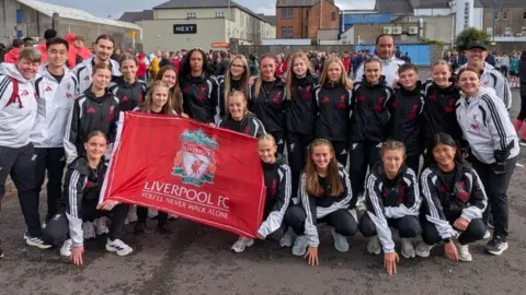 A team of young footballers are assembled for a group photo. They are wearing their team tracksuit and have their arms around each other. A number of them are smiling. 