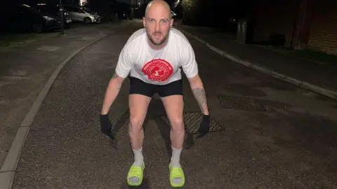 Daniel Fairbrother Daniel Fairbrother wearing bright green slider shoes on his feet, with white socks, crouching down, to show them, in a street, wearing a T-shirt, black shorts and black gloves. It is dark and you can just see cars and houses. 