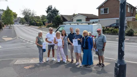 Oliver Jacques A group of around nine people stand together on a pavement beside a road, several holding printed flyers. The wide residential street stretches into the distance behind them with houses on both sides. They are standing near a lamppost on what appears to be a sunny day.