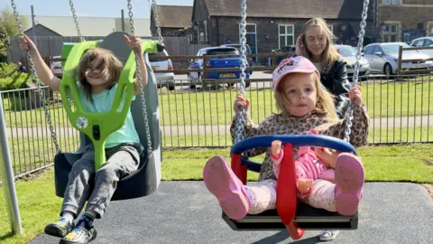 North Northamptonshire Council Two children in swings in a play area, with a woman behind them and houses and cars behind her. One child is wearing a pink cap and the other a T-shirt and trousers. 