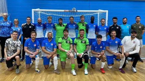 Steve Down A team photo of the England men's deaf futsal team. They are standing in front of a goal inside an indoor sports hall. They are wearing a blue and white kit, although the goalkeepers are wearing green.