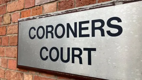 BBC A silver plaque on the outside of the court. The words 'coroners court' are written in bold capital letters in black. The sign is on a brick wall. 