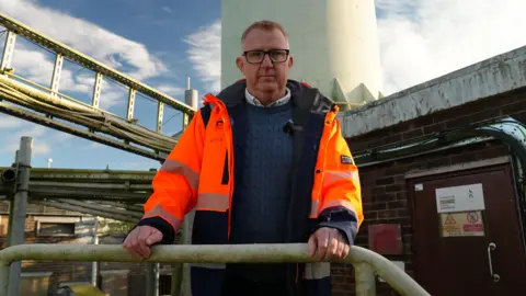 A man wearing a bright orange high‑visibility jacket stands on a metal platform, holding onto a handrail with both hands. Behind them rises a tall, pale structure. Metal pipes and framework stretch across the background to the left, and a brick building with warning signs on its door sits to the right. The sky above is clear with scattered clouds.