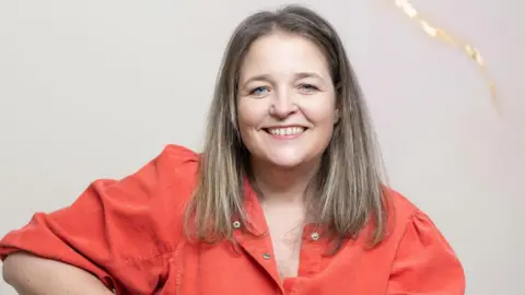 A woman with straight blonde hair and an dark orange shirt is smiling at the camera against a plain background