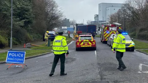 Two police officers standing at a junction of a road. There is a blue police sign to the left of the image and there is a fire car, a police car and a fire engine in front of them.