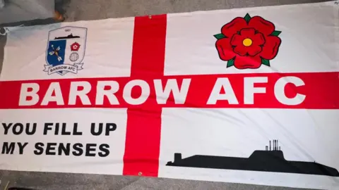 Supplied A large England flag with the words Barrow AFC across the horizontal line of the red cross. In the top left corner it has the Barrow AFC badge, the top right has a red rose, the bottom left has the phrase "you fill up my senses" and the remaining quarter has the outline of a black submarine. 