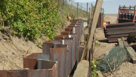 John Fairhall/BBC A close-up of sheet metal barriers on the left after they have been installed where a cliff meets a beach. They are rusty brown coloured and have been installed in a zig-zag shape. Beyond them on the right is the beach, with some blue fishing net, and piles of more sheet metal barriers and beyond that the sea and sky.