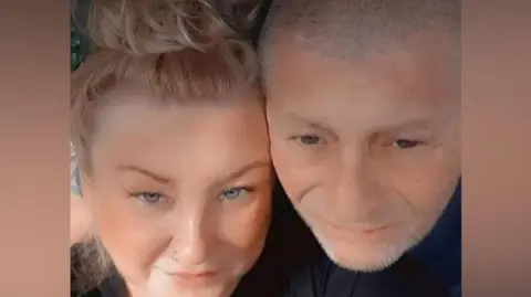 Suzanne Hall A selfie showing Suzanne Hall, who has blonde hair pulled back into a ponytail, cheek to cheek with her husband, who had short grey hair and grey stubble