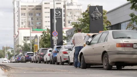 BBC A man in a white t-shirt facing away from the camera stands with his right arm resting on his car, in a long queue of vehicles outside a petrol station in Moscow