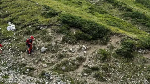 Salvamont Brașov A man wearing a red hat, black shirt and red trousers, with a red backpack and black walking stick, walks along a rocky hillside.