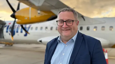 Paul Saunders wearing a blue shirt and blue suit jacket and black glasses. He is stood in front of an Aurigny aircraft which is blurred. 