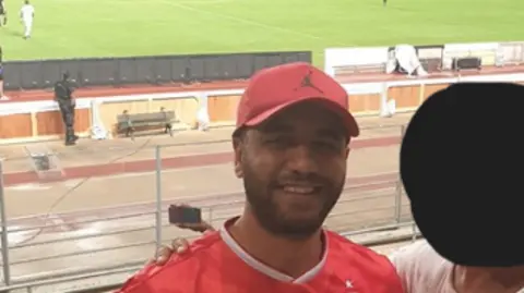 Department for Work and Pensions Adam Mejri stands in a stadium posing for a photo as a cricket match takes place in the background. He wears a red shirt and a red cap.