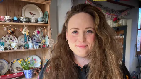 Phoebe Day has long brown hair and blue eyes. She is standing in front of a wooden piece of furniture with decorative china on its shelves.