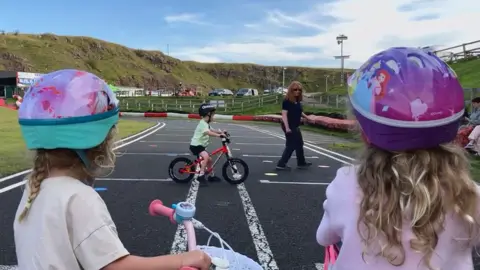 Two young girls on bikes facing away from the camera and wearing cycle helmets, watching a young boy being coached in learning to ride. The coach, Fiona Graham, is wearing a blue polo shirt and trousers and is walking near the boy.