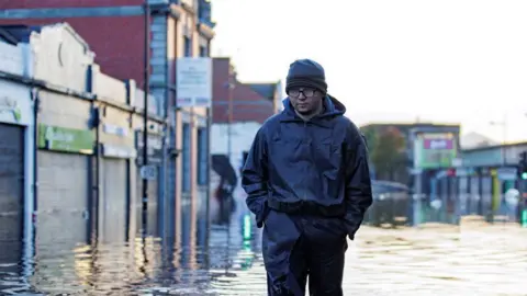 a man in waterproofs and welly boots walking through water that is up to his shins. On either side of him, shopfronts are visible, making it clear he is walking down a street