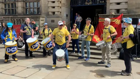 A group of people wearing yellow t-shirts are playing drums in the street outside Sheffield town hall