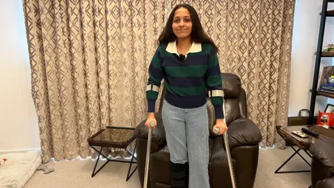A 12-year-old girl wearing a navy and green rugby shirt and light blue jeans smiles as she stands using crutches in her living room. There is a brown leather armchair behind her.