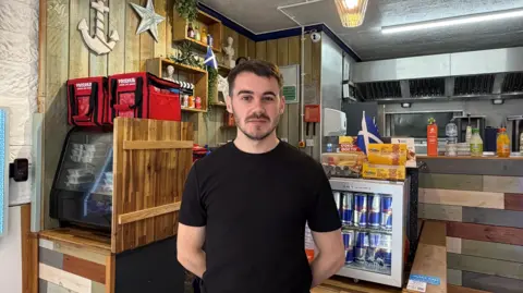 A man wearing a black t-shirt stands in front of a food preparation area. There are Scottish flags and food storage containers behind him.
